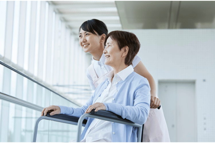 窓を見つめる高齢者で車椅子の女性を女性介護士がケアする様子